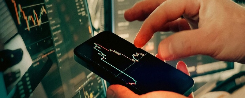 스마트폰으로 실시간 주식 시장 그래프와 경제 데이터를 분석하고 있는 전문가의 손과 사무실 배경