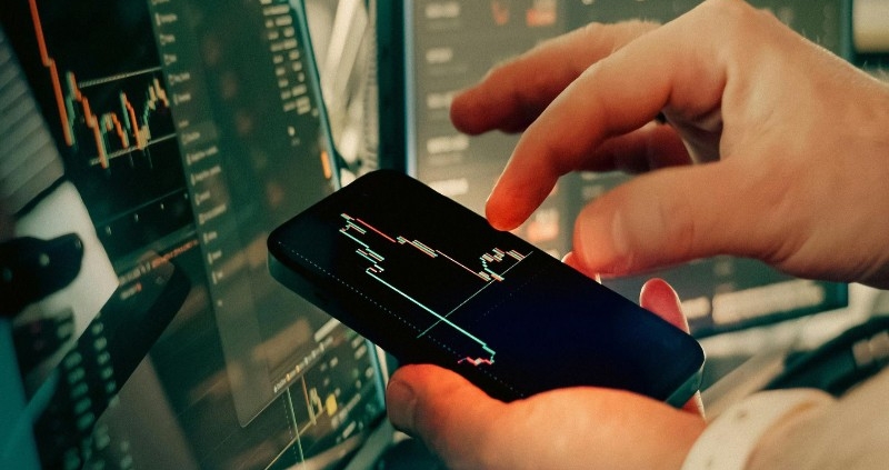 스마트폰으로 실시간 주식 시장 그래프와 경제 데이터를 분석하고 있는 전문가의 손과 사무실 배경