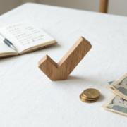 Wooden checkmark symbol on a white tablecloth near coins, Japanese yen bills, and an open notebook with handwritten notes.